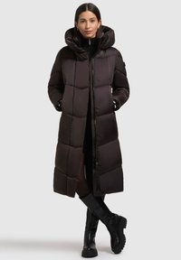 Pitkä musta toppatakki hupulla, jossa on tikattu kuosi, edessä vetoketju ja napit, sekä sivutaskut. Päällä mustat vaatteet.