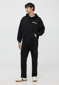 Felpa nera con tasca frontale, testo "DARKER" in bianco, abbinata a pantaloni cargo neri con tasche laterali e scarpe da ginnastica bianche.