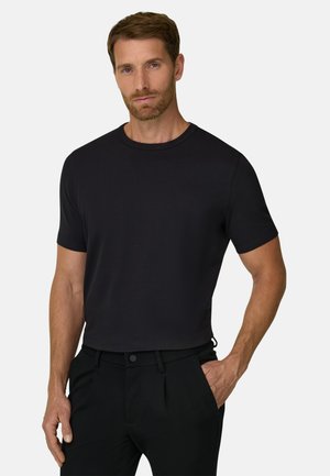 Mand med kort brunt hår og skæg iført en ensfarvet sort kortærmet t-shirt og sorte bukser med den ene hånd i lommen.
