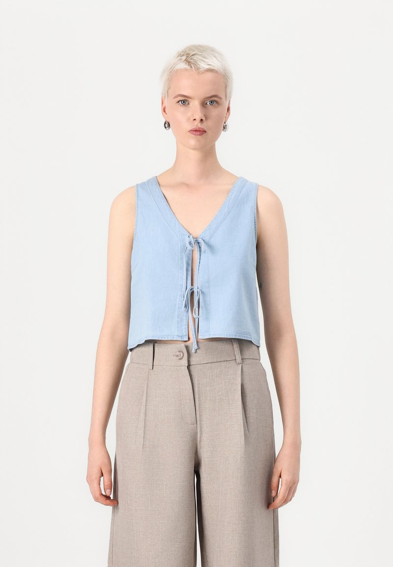 Lichtblauw cropped top met een diepe V-hals, geknoopte voorkant en brede bandjes, gecombineerd met beige op maat gemaakte high-waisted broeken.