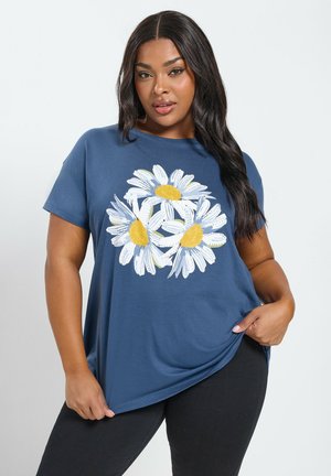 Frau mit langen dunklen Haaren, die ein blaues T-Shirt mit drei großen weißen und gelben Gänseblümchen trägt und mit einer Hand den Saum des T-Shirts hält.