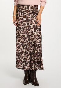 Vrouw in een satijnen midi rok met luipaardprint, roze trui, zilveren armbanden, ringen en donkerbruine gehuurde enkelboots tegen een witte achtergrond.