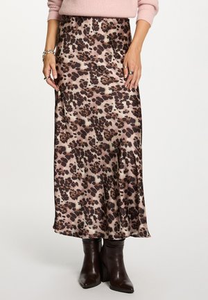 Vrouw in een satijnen midi rok met luipaardprint, roze trui, zilveren armbanden, ringen en donkerbruine gehuurde enkelboots tegen een witte achtergrond.