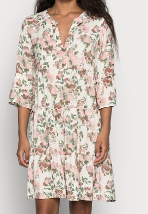 Vrouw draagt een losse, witte jurk met bloemen in roze en groen, driekwart mouwen en een V-hals.