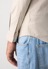 Beige overhemd met lange mouwen, manchet detail boven de lichtblauwe denim jeanszak, met een zichtbare kleine merklabel op de zijnnaad van het shirt.