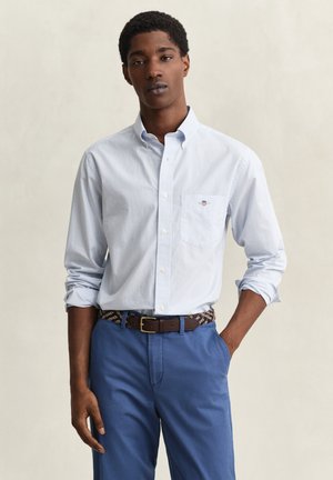 Junger Mann, der ein hellblau kariertes, knöpfbares Hemd mit Brusttasche und hochgekrempelten Ärmeln trägt, blaue Hosen und einen gewebten Gürtel, stehend mit einer Hand in der Tasche.