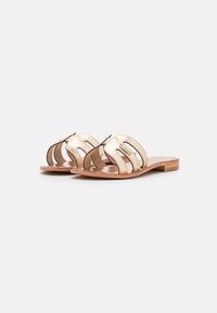 Sandali in pelle dorata con suole di legno piatto, caratterizzati da motivi ritagliati e dettagli cuciti, che offrono uno stile open-toe e un design minimalista.