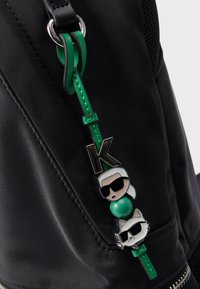 Juodos nailono kuprinės detalė su žaliu dirželiu ir trimis dekoratyviniais pakabukais: raide „K“, figūra su saulės akiniais ir kate.