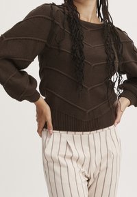 Brown knitted sweater with textured chevron pattern, ribbed cuffs, and hem. Paired with cream pants featuring vertical burgundy stripes.