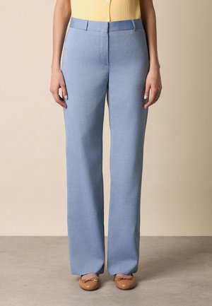 Vrouw in lichtblauwe rechte broek, gele mouwloze top met knopen en beige ballerina's, staand op een grijze vloer.