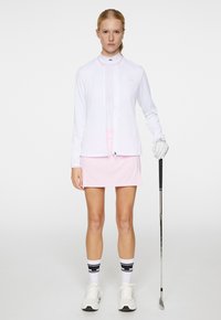 Vit ribbad långärmad jacka med en rosa detalj, över en vit åtsittande skjorta och rosa skorts, bärs med vita skor och svarta randiga strumpor.