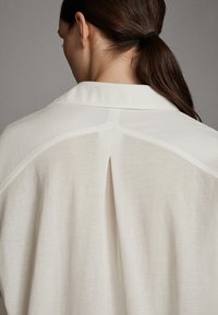 Ljusbeige blus med en bakfick, mjuk textur och klassisk krage. Designad för en avslappnad passform med minimala sömnadsdetaljer.