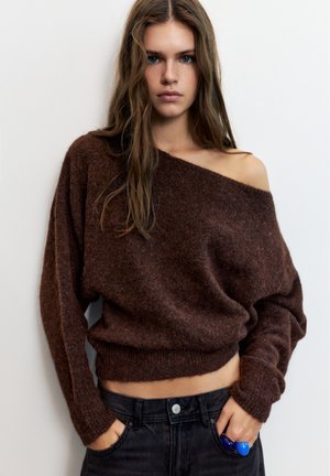 Jeune femme aux longs cheveux portant un pull marron décolleté aux épaules et un jean noir, mains dans les poches, sur un fond blanc uni.