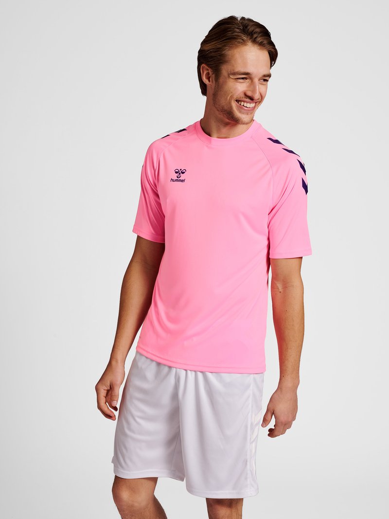 Hombre sonriente con una camiseta deportiva rosa brillante con chevrones azul marino en los hombros y pantalones cortos deportivos blancos, de pie contra un fondo liso.