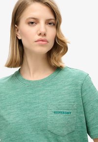 Grøn bomulds T-shirt med rund hals, korte ærmer og en lomme. "SUPERDRY" er broderet i en kontrastfarvet teal på lommen.
