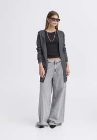 Sivi dolg pulover čez črn kratki top, kombiniran z sivimi širokimi kavbojkami. Obleka je sestavljena iz enobarvnih in minimalističnih oblik.