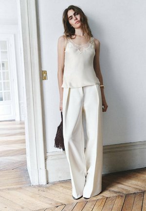 Femme debout portant un haut blanc sans manches orné de dentelle et un pantalon large crème, tenant un sac à main marron foncé à franges.