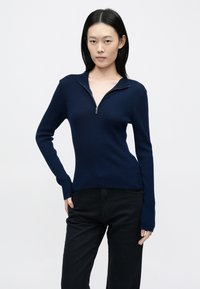 Tengerészkék bordázott kötött pulóver cipzáras gallérral és hosszú ujjakkal, fekete, magas derekú nadrággal párosítva. Simaságú, testhez álló fazon.