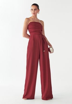 Czerwony kombinezon bez ramiączek, z zaznaczoną talią i szerokimi nogawkami. Wykonany z gładkiej tkaniny z minimalnymi ozdobami.
