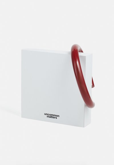 Bolsa rectangular blanca con un asa roja brillante. Presenta el texto de la marca "uncommon matters" en la parte frontal. Diseño simple y moderno.