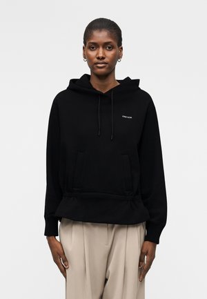 Kvinde iført sort Stine Goya hoodie med forlomme og beige plisserede bukser, stående mod en enkel lys baggrund.
