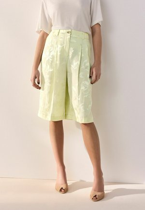 Kobieta nosząca bladoróżowe kwieciste szorty o szerokich nogawkach sięgające kolan, w połączeniu z białą bluzką i beżowymi sandałami na otwartym palcu, stojąca na tle jednolitej ściany.