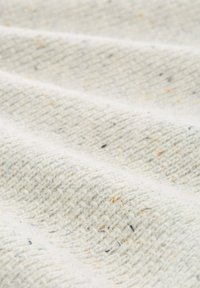 Textielstof in crème met een geweven textuur, voorzien van kleine vlekjes in oranje en zwart door het materiaal heen. Gekreukt en gelaagd uiterlijk.