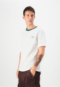 Camiseta de algodón blanca con un cuello de canalé verde y un pequeño logo bordado. Presenta mangas cortas y un corte relajado.