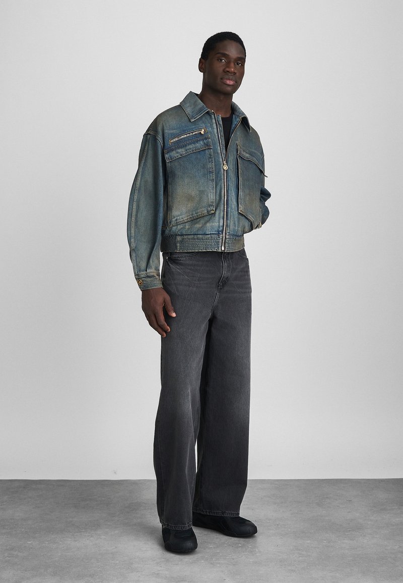 Jeansjacke in verblichendem Blau mit zwei Brusttaschen und einem Reißverschluss. Kombiniert mit locker sitzenden grauen Jeanshosen und schwarzen Schuhen.