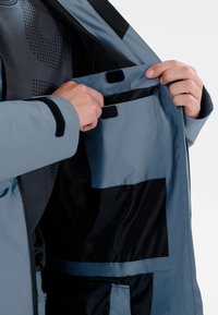 Blå vandtæt jakke indvendig med en velcrolomme, sort foring og tekstureret gråt stof nedenunder, designet til funktionalitet og bekvemmelighed.