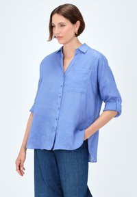 Camicia button-up in lino blu con una sola tasca sul petto, maniche arrotolate e una vestibilità rilassata, abbinata a pantaloni in denim blu scuro.