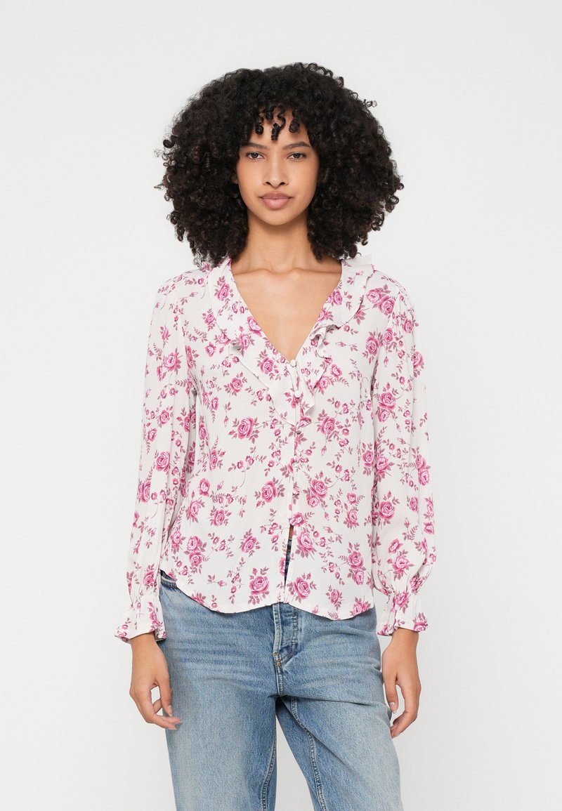 Blusa floreale in bianco con motivi di rose rosa, con scollatura a V e chiusura a bottoni. Le maniche lunghe hanno polsini arricciati. Tessuto morbido.
