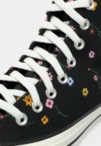 Zwarte canvas sneaker met witte veters, voorzien van kleurrijke bloemenpatronen in roze, geel en blauw. Zilveren oogjes en een witte rubberen zool.