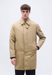 Νεαρός άνδρας που φοράει μπεζ μονόμπαστο καμπαρντίνα με γιακά, όρθιος με το ένα χέρι στην τσέπη σε απλό λευκό φόντο.