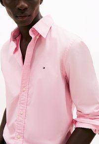 Lys rosa skjorte i bomull med knapper, krage, oppbrettede ermer og en liten logo på brystet. Glatt tekstur.