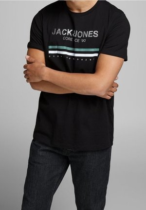 Man wearing black Jack & Jones t-shirt with green and white stripes, arms crossed, paired with dark jeans against plain background.