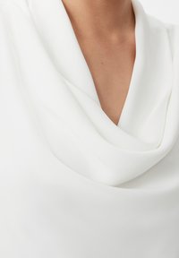 Blusa bianca con collo a cowl realizzata in tessuto liscio. Presenta un design drappeggiato e una texture morbida, con una scollatura profonda e dettagli di cucitura minimi.