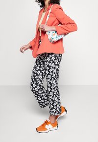 Korallfärgad kavaj, gul topp, svarta blommiga byxor och orange sneakers. Handväskan har en vit bas med ett färgglatt mönster av frukter och löv.