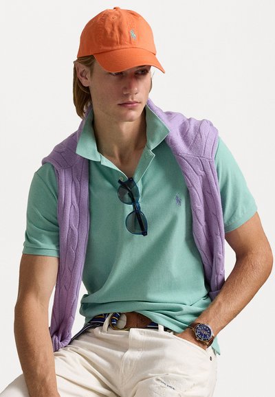 Polo shirt in mintgroen met een kraag, lavendel kabelgebreide trui over de schouders, oranje pet, donkere zonnebril en witte broek.