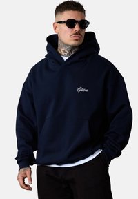 Hoodie surdimensionné bleu marine avec une poche avant et un logo brodé. Présente une capuche avec cordon de serrage et des poignets côtelés pour un ajustement confortable.