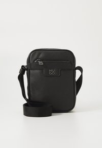 Sort læder crossbody taske med struktureret overflade, lynlåslomme foran og justerbar rem. Logo detalje på fronten. Rektangulær form.