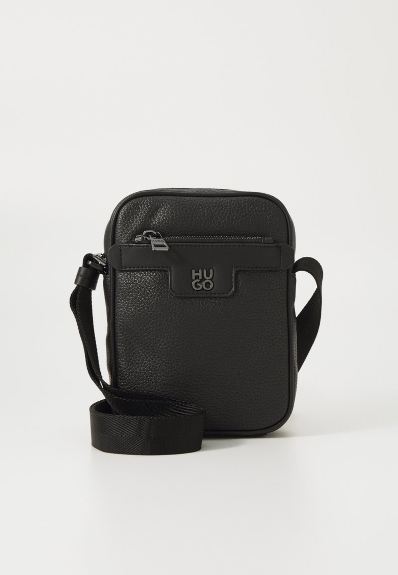 Sort læder crossbody taske med struktureret overflade, lynlåslomme foran og justerbar rem. Logo detalje på fronten. Rektangulær form.