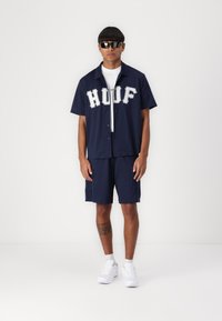 Marineblauw overhemd met korte mouwen en geperforeerde textuur, met de letters "HDUF", gecombineerd met marineblauwe shorts. Gemaakt met een zonnebril en witte sneakers.