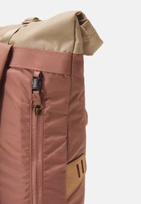 Rolled-top backpack in a two-tone design: pink fabric with a matte finish and beige top. Features a side zip pocket and cork accent strap.
