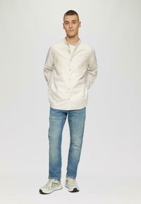 Wit en beige gestreept overhemd, dichtgeknoopt, met een bandkraag; lichtblauwe spijkerbroek; grijze sportschoenen. Effen achtergrond.