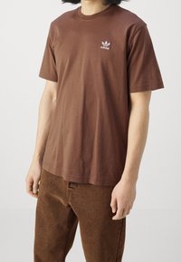 Hnědé bavlněné tričko s krátkým rukávem, kulatým výstřihem a malým bílým logem Adidas na levé hrudi. Nosené s hnědými texturovanými kalhotami.