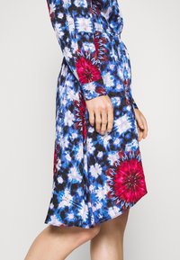 Robe en coton avec un imprimé floral bleu, noir et rouge. Manches longues avec poignets à boutons et taille élastique pour une silhouette ajustée.