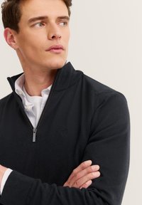 Junger Mann mit verschränkten Armen, der einen schwarzen Reißverschluss-Pullover über einem weißen Hemd trägt und seitlich gegen einen schlichten Hintergrund schaut.