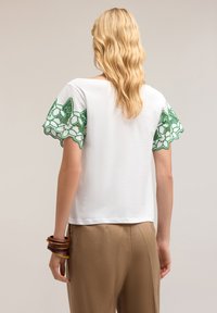 Top corto bianco a maniche corte con motivi floreali ricamati in verde sulle maniche. Tessuto morbido, vestibilità rilassata, abbinato a pantaloni marroni.