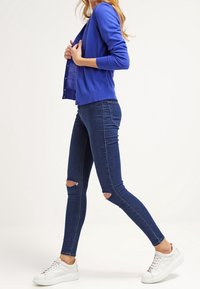 Modrý kardigan s knoflíkovými detaily, nošený přes přiléhavý bílý top, doplněný tmavě modr�ými roztrhanými úzkými džíny a bílými teniskami.